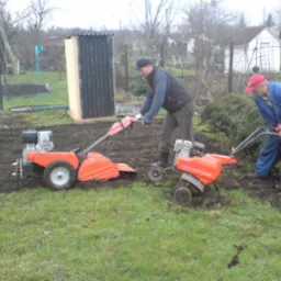 Dwaj mężczyźni pracują glebogryzarkami spalinowymi na działce: jeden przygotowuje ziemię pod uprawy, drugi obok krzewów, w tle widoczna budka i dom.