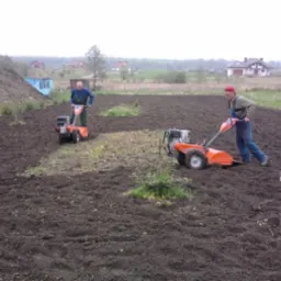 Dwaj mężczyźni używają glebogryzarek spalinowych do przekopywania ziemi na działce w pochmurny dzień. Widoczne domy w tle.