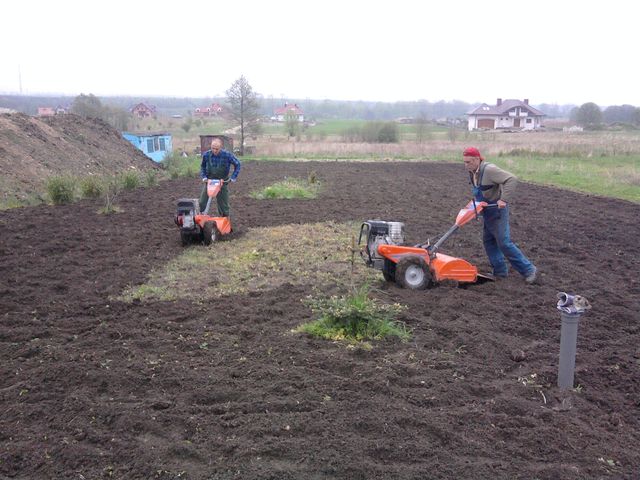 Dwaj mężczyźni używają glebogryzarek spalinowych do przekopywania ziemi na działce w pochmurny dzień. Widoczne domy w tle.
