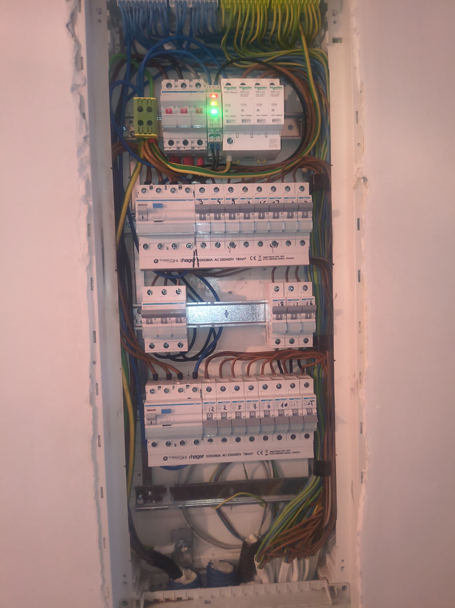 Rozdzielnica elektryczna Hager z zainstalowanymi bezpiecznikami i przewodami w kolorach: niebieskim, brązowym, zielono-żółtym. Widoczne oznaczenia na urządzeniach.