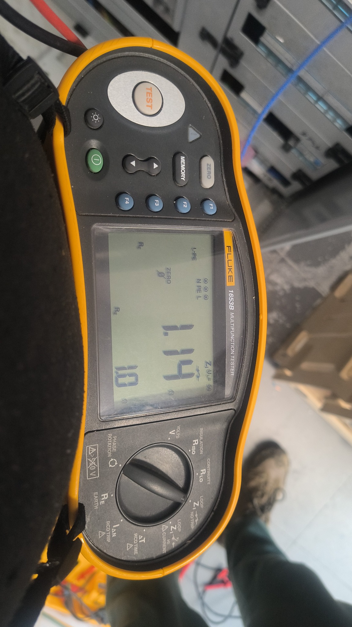 Żółty miernik Fluke 1653B z wyświetlaczem wskazującym 1.14 Ω, używany do pomiarów elektrycznych w szafie serwerowej. Widoczne kable i fragment obuwia.