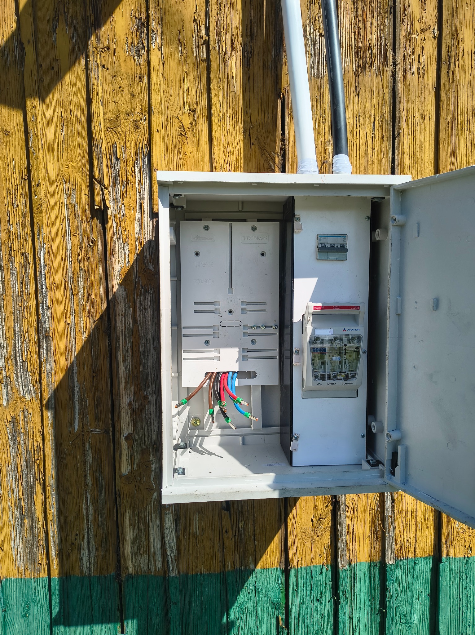 Otwarta skrzynka elektryczna z licznikiem i bezpiecznikami, zamontowana na żółto-zielonej, drewnianej ścianie. Widoczne kable i elementy instalacji.