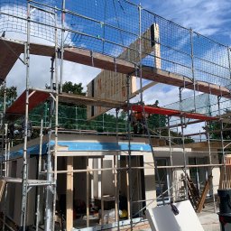 ARCHIQUALITY Krystian Nowak - Montaż ściany domu szkieletowego w Dębnie przy użyciu dźwigu, widoczne rusztowania i pracownicy budowlani.