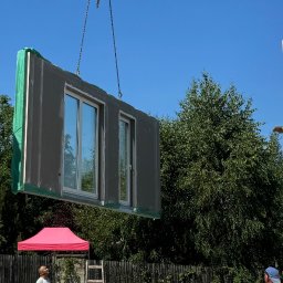 ARCHIQUALITY Krystian Nowak - Montaż modułu domu z oknami za pomocą dźwigu, widoczni dwaj pracownicy, drabina oparta o ogrodzenie, w tle zieleń drzew.