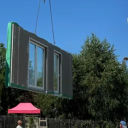 Montaż modułu domu z oknami za pomocą dźwigu, widoczni dwaj pracownicy, drabina oparta o ogrodzenie, w tle zieleń drzew.