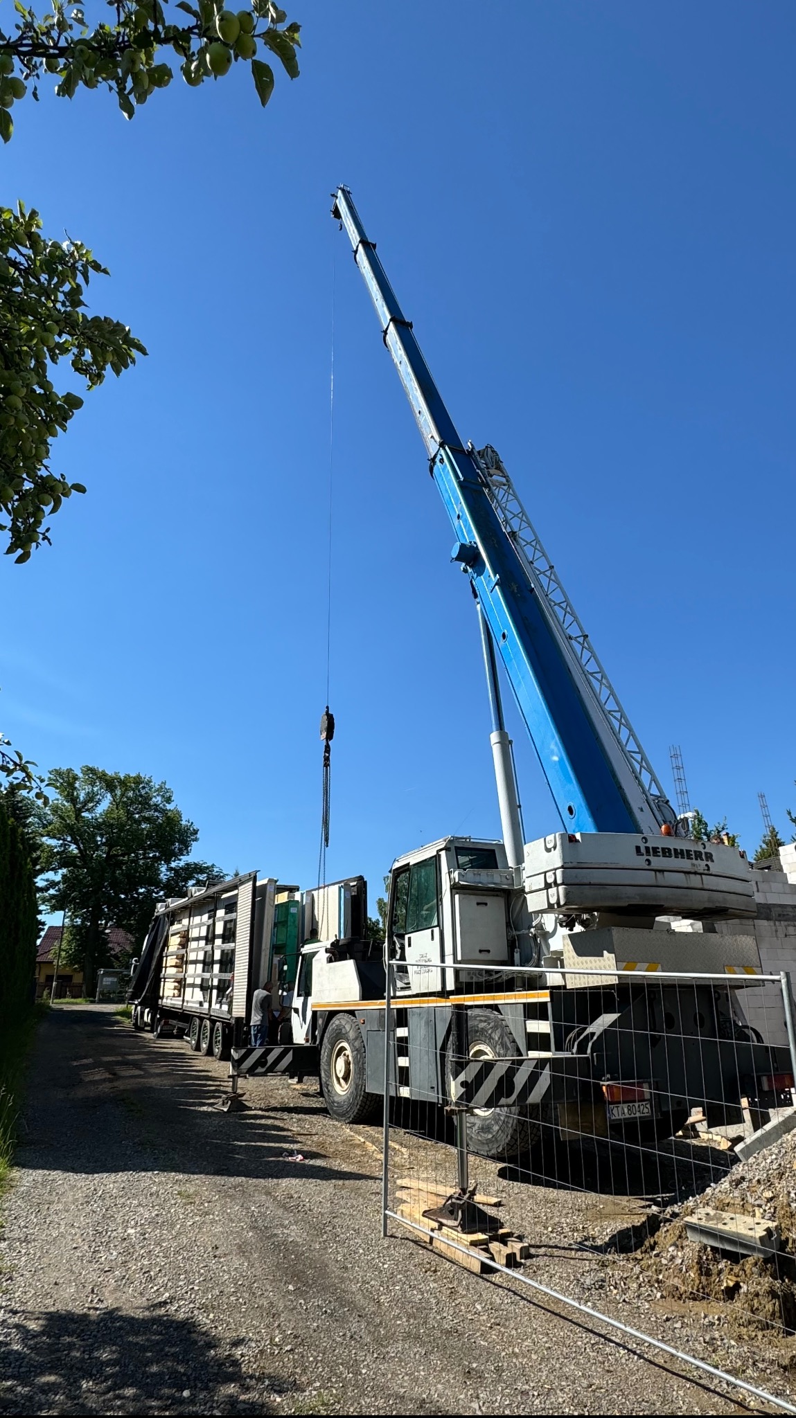 Żuraw Liebherr podnosi element konstrukcyjny domu, widoczne elementy domu na lawecie, ogrodzenie placu budowy.