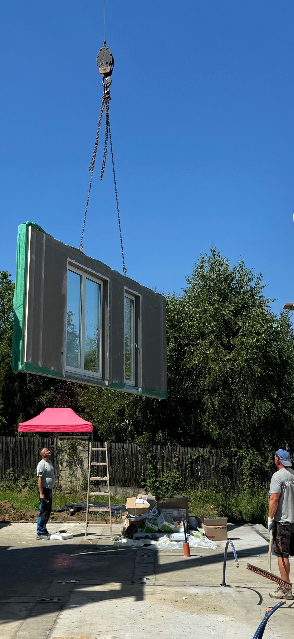 Montaż modułu domu z oknami za pomocą dźwigu, widoczni dwaj pracownicy, drabina oparta o ogrodzenie, w tle zieleń drzew.