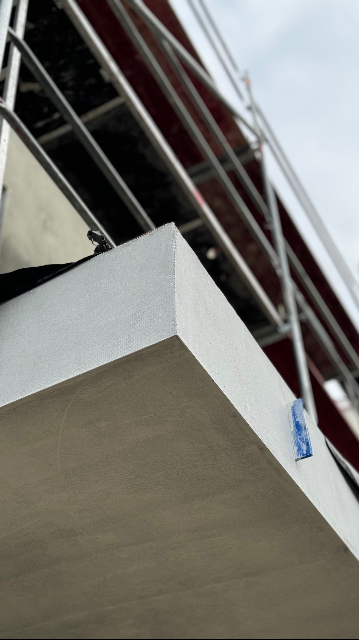 Fragment ściany budynku w trakcie budowy, widoczne rusztowanie i niebieski element mocujący.