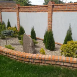 Ogródek przed domem z niskim murkiem z cegły, wysypany kamykami, z ozdobnymi krzewami i kamieniem na tle muru z ceglanymi słupkami i białym wypełnieniem.