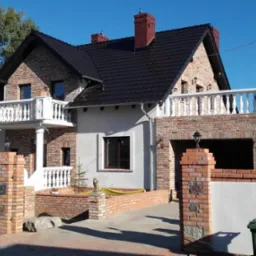 Dwukondygnacyjny dom z cegły klinkierowej z balkonami z białymi balustradami, czarnym dachem i garażem, otoczony niskim murem z cegły i białymi panelami.