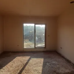 Wnętrze pokoju po nałożeniu świeżego, jasnobeżowego tynku na ścianach, z widocznym oknem i niedokończoną podłogą w stanie surowym.