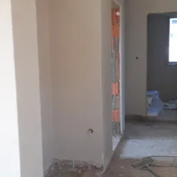 Wnętrze w trakcie prac wykończeniowych: świeżo otynkowane ściany w odcieniach beżu, widoczne surowe cegły w otworze drzwiowym, niezagruntowana podłoga i wystające kable elektryczne.