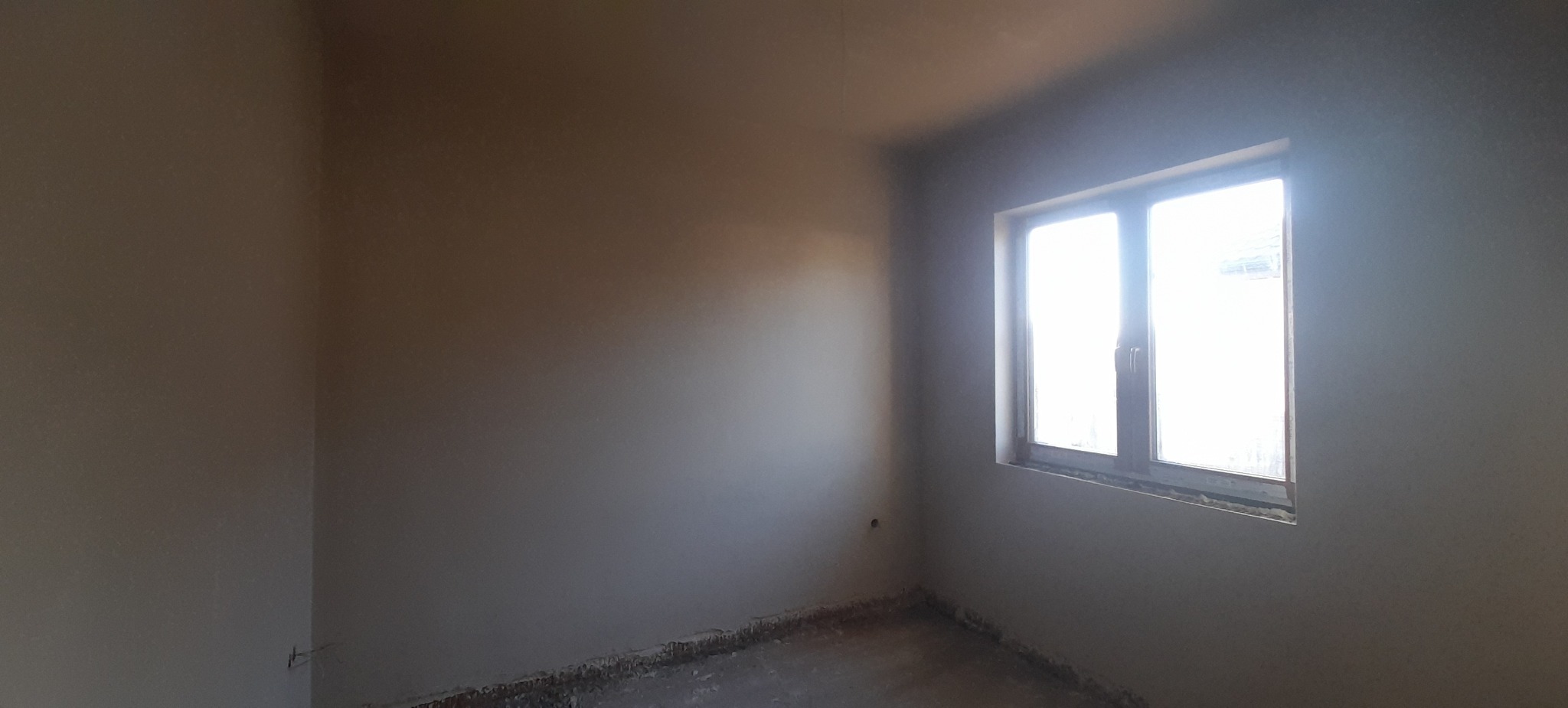 Pokój po nałożeniu gładzi szpachlowej, widoczne ślady po pracach wykończeniowych na podłodze, okno z ramą drewnianą wpuszczające jasne światło.