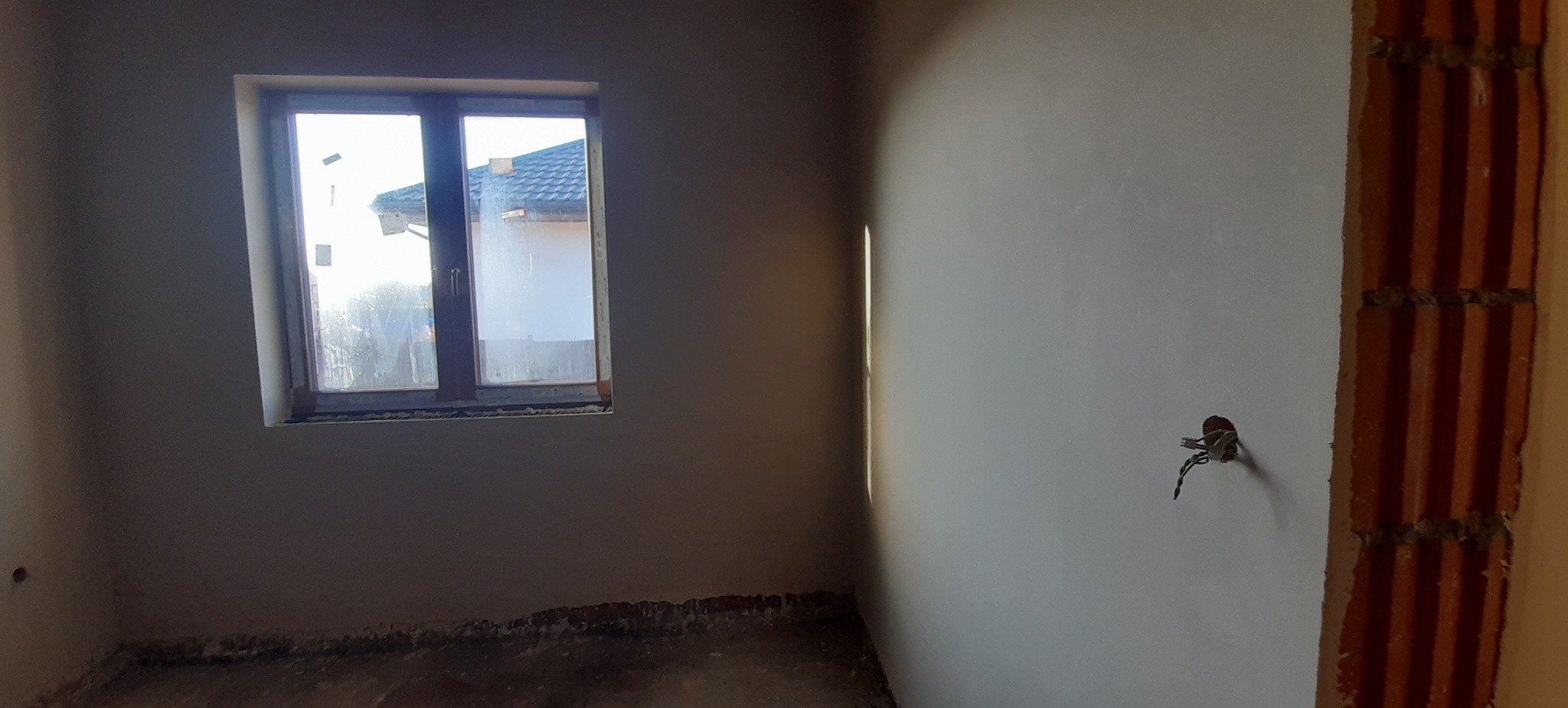 Świeżo otynkowane wnętrze z widoczną instalacją elektryczną i fragmentem ściany z cegły dziurawki, widok przez brudne okno na dom sąsiada.