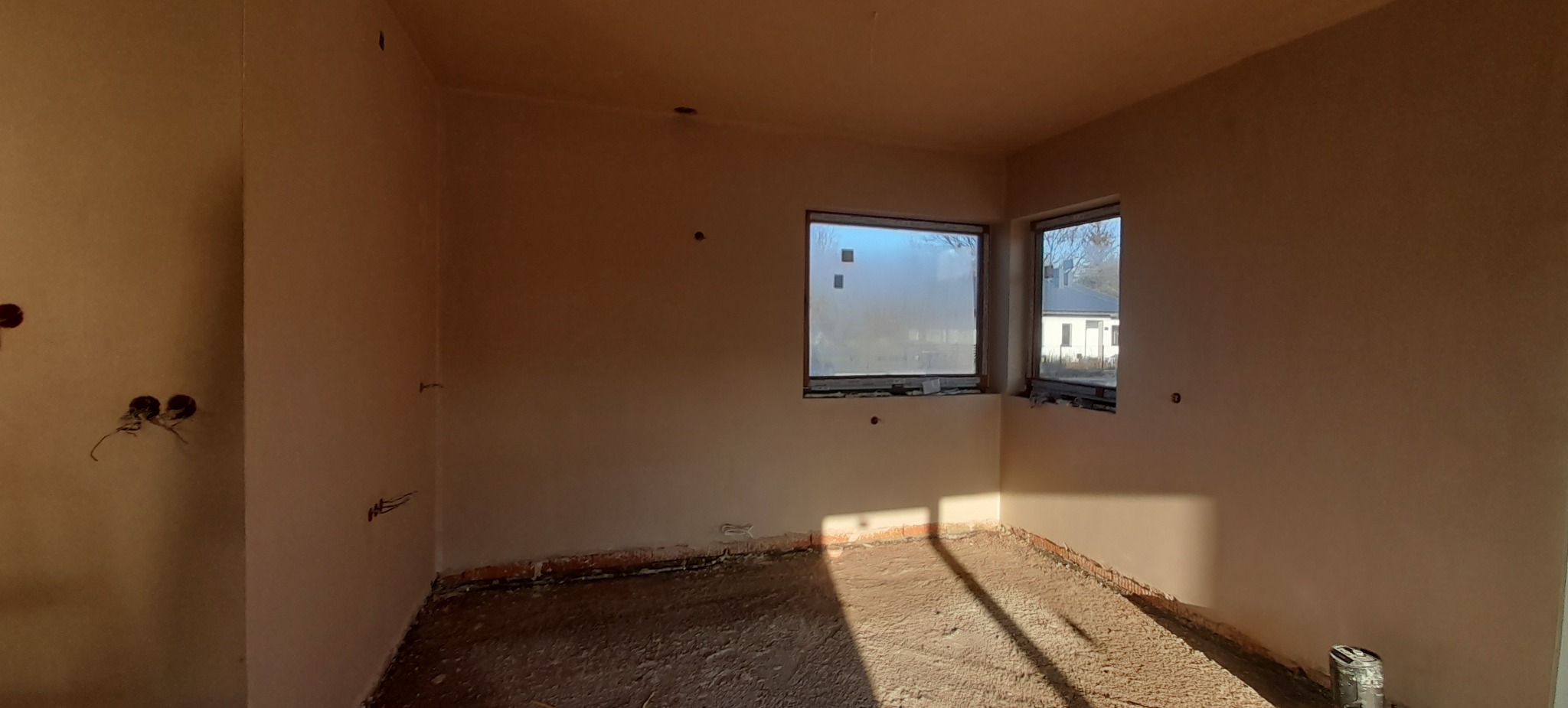 Świeżo otynkowane wnętrze z dwoma oknami i widocznymi puszkami elektrycznymi, surowy stan podłoża.