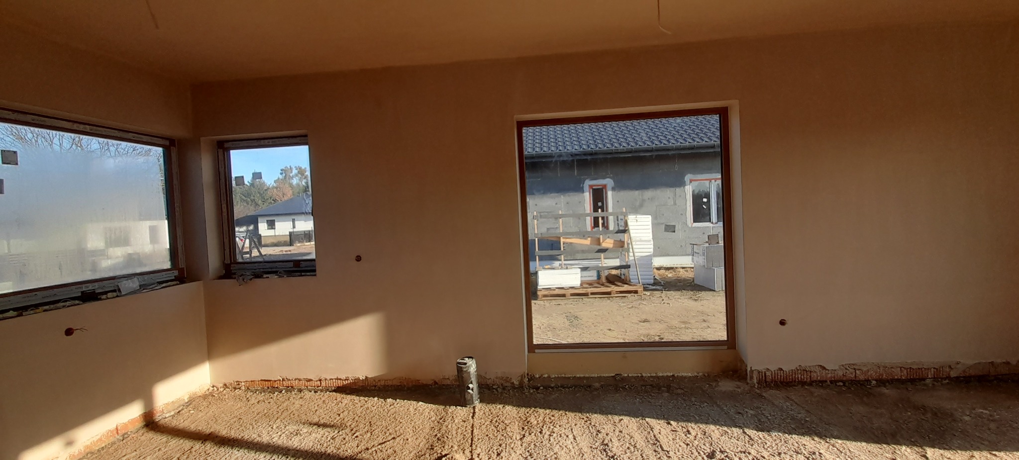 Wnętrze w stanie deweloperskim z nałożonymi jasnymi tynkami na ścianach, widoczne okna z widokiem na budynki na zewnątrz, podłoga z pozostałościami po pracach budowlanych.