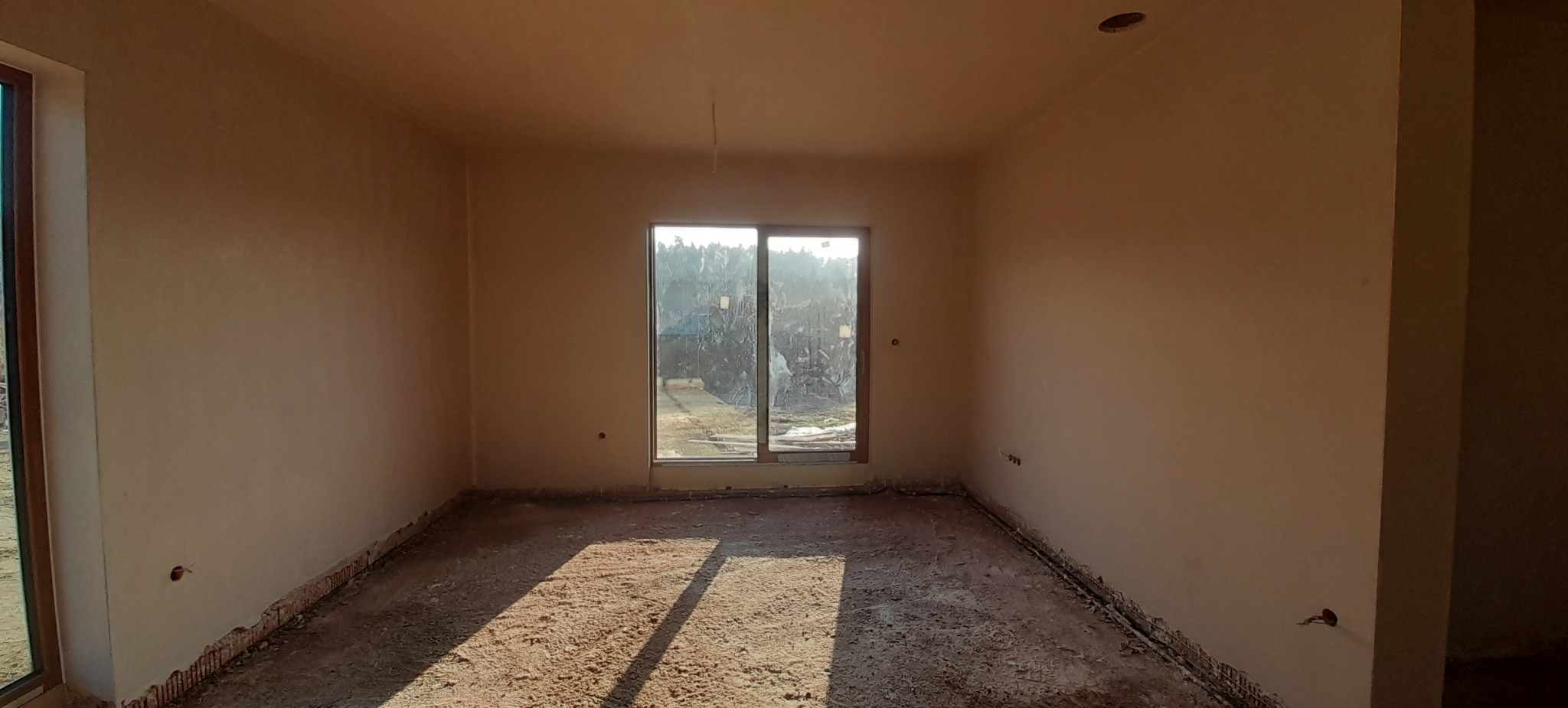 Wnętrze pokoju po nałożeniu świeżego, jasnobeżowego tynku na ścianach, z widocznym oknem i niedokończoną podłogą w stanie surowym.