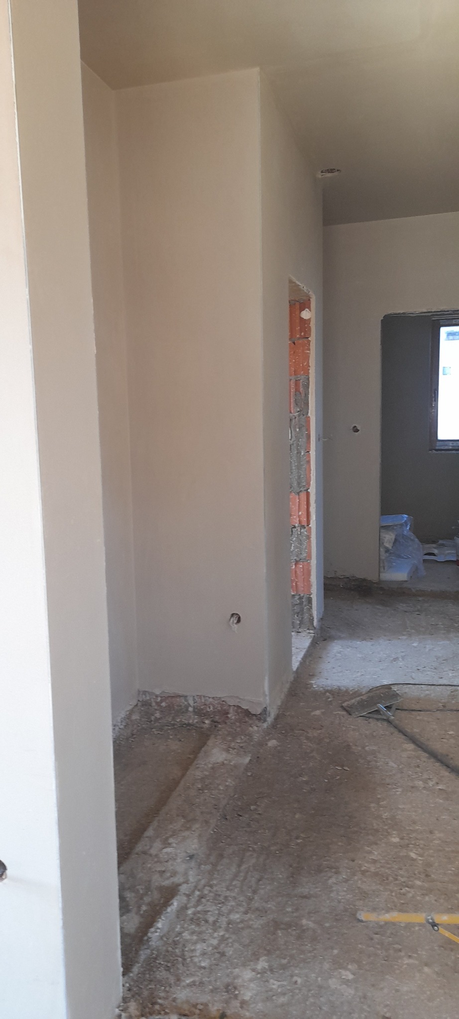 Wnętrze w trakcie prac wykończeniowych: świeżo otynkowane ściany w odcieniach beżu, widoczne surowe cegły w otworze drzwiowym, niezagruntowana podłoga i wystające kable elektryczne.