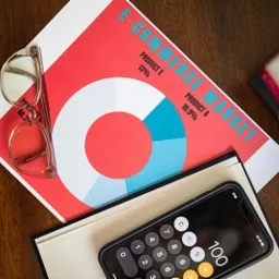 Analiza rynku e-commerce na wykresie kołowym z kalkulatorem na smartfonie, notatnikiem i okularami w złotych oprawkach.