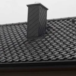 Czarny dach z dachówki ceramicznej z kominem obłożonym łupkiem, widoczne rynny i odpowietrznik.