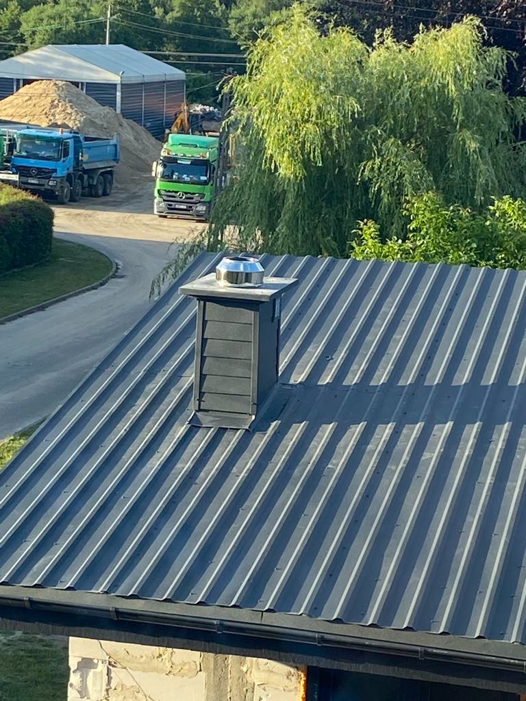 Nowoczesny dach z blachy trapezowej w kolorze grafitowym, z kominem wentylacyjnym. W tle widoczne ciężarówki i hałda piasku, co sugeruje trwające prace budowlane.