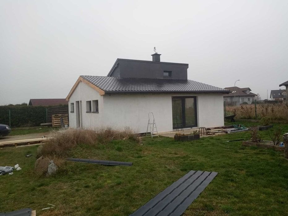 Dom jednorodzinny z nowym, ciemnym dachem z dachówki ceramicznej, widoczne elementy więźby dachowej i drabina oparta o ścianę, otoczenie w trakcie prac wykończeniowych.