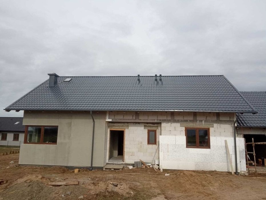 Dom w trakcie budowy z zamontowanym grafitowym dachem z blachodachówki modułowej, widoczne rynny i komin. Część budynku otynkowana, druga w surowym stanie z białego bloczka.
