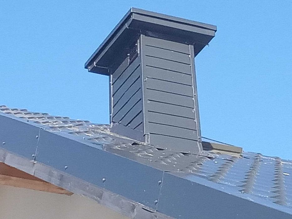 Szary komin z metalowym wykończeniem na dachu z blachy trapezowej, widoczny na tle błękitnego nieba.