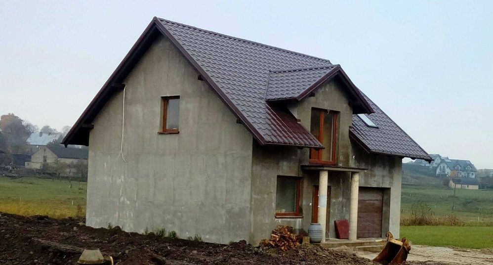 Surowy dom jednorodzinny z brązowym dachem z blachodachówki, jeszcze bez elewacji, widoczne rynny, okna i drzwi w brązowych ramach, na pierwszym planie ziemia i gruz.