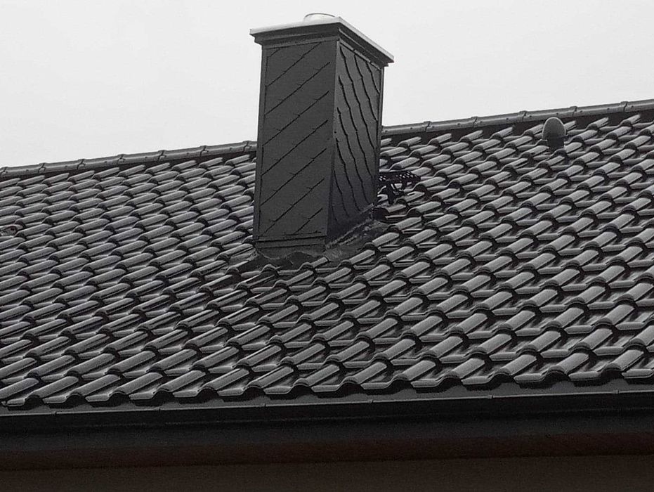 Czarny dach z dachówki ceramicznej z kominem obłożonym łupkiem, widoczne rynny i odpowietrznik.