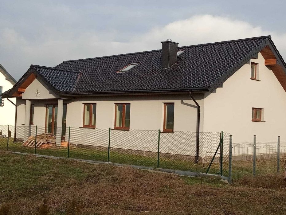 Dom jednorodzinny z nowym, ciemnym dachem z dachówki ceramicznej, widoczne rynny, komin i okna dachowe, ogrodzony siatką.