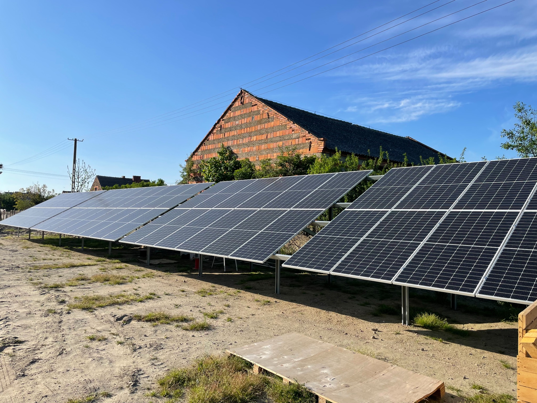 Rząd paneli słonecznych na polnej działce przed ceglanym budynkiem w słoneczny dzień.