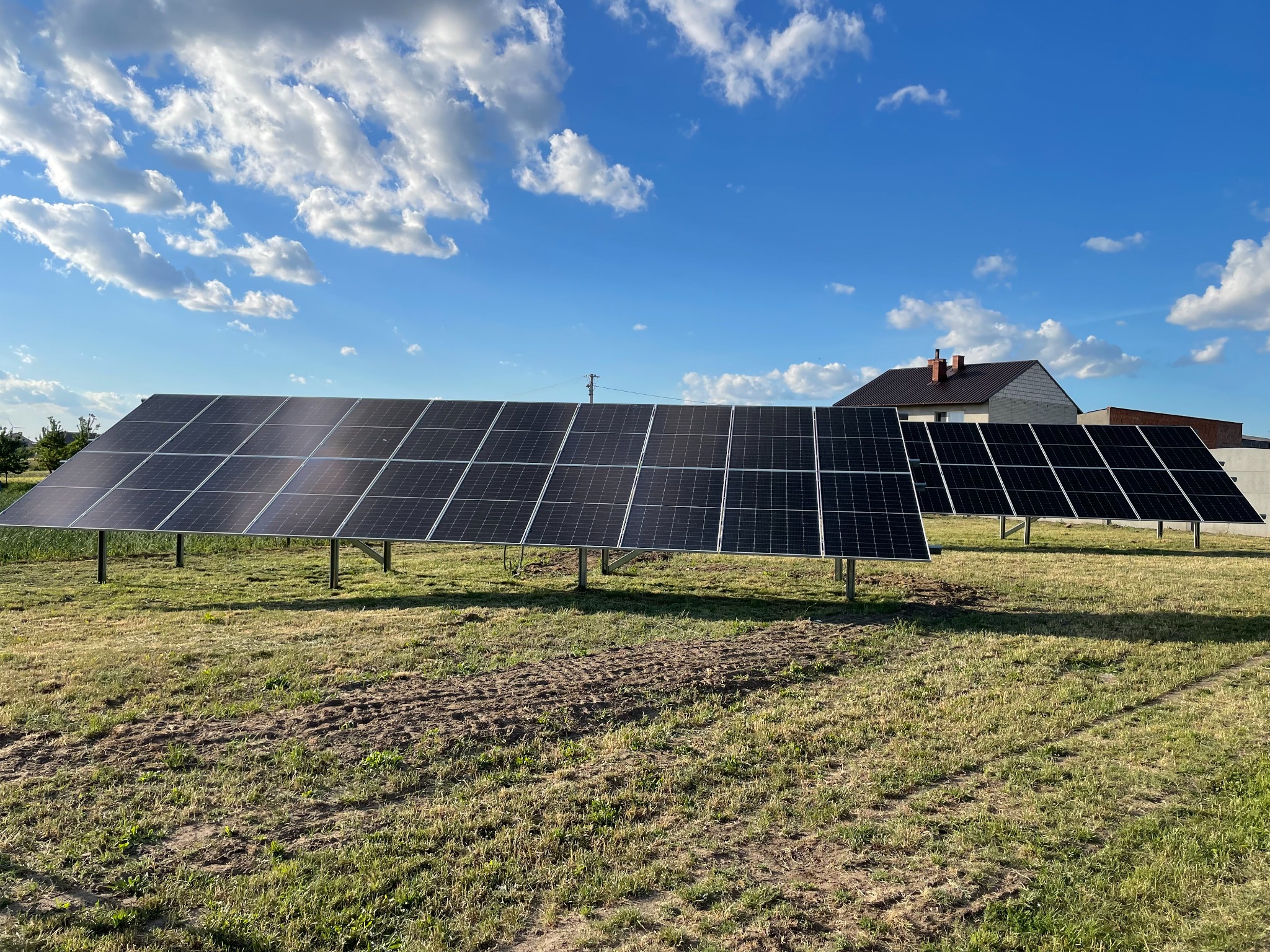 Dwa rzędy paneli słonecznych na metalowych konstrukcjach, ustawione na trawiastym polu pod błękitnym niebem z białymi chmurami. W tle widoczny budynek z brązowym dachem.