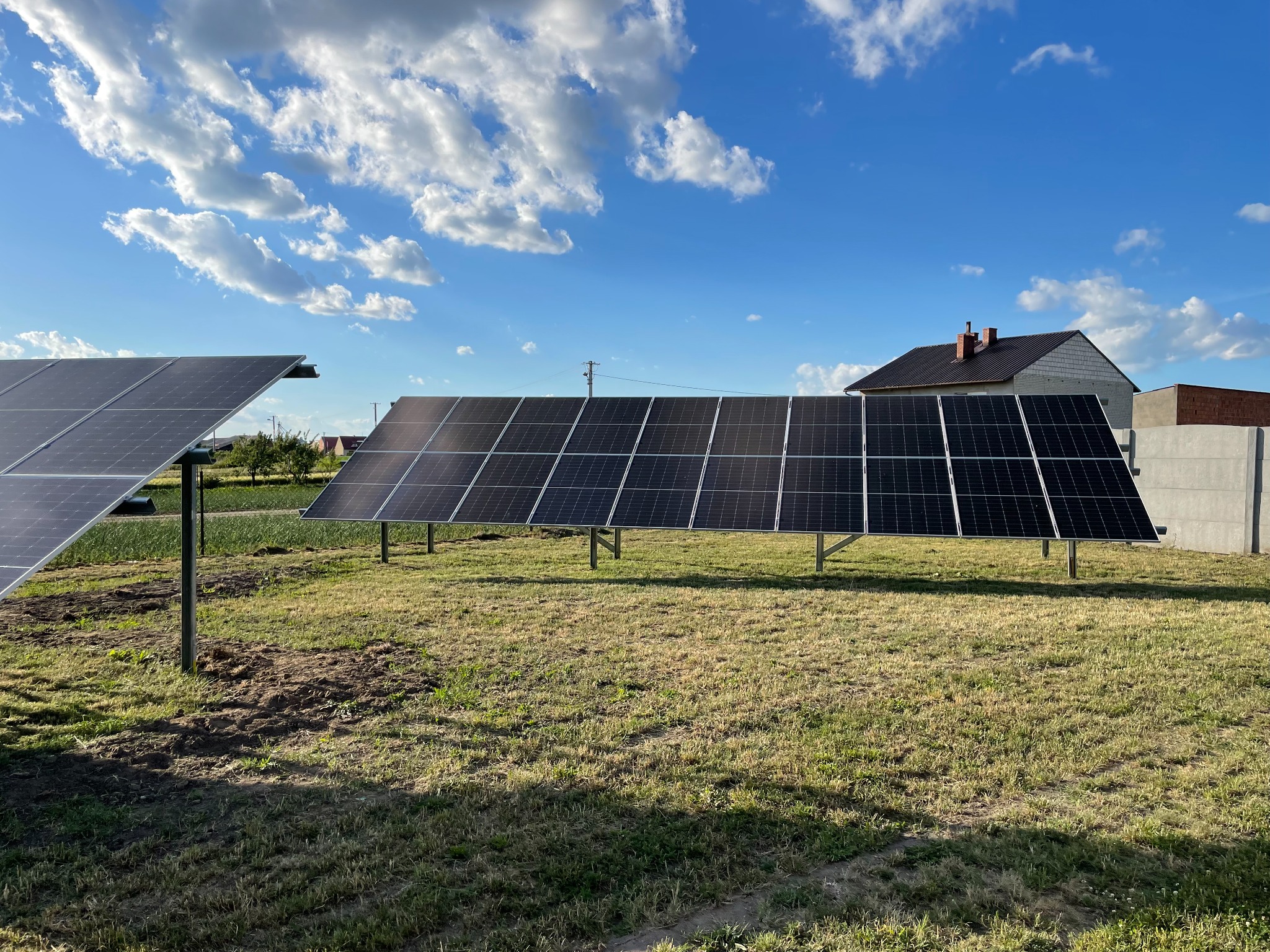 Instalacja paneli słonecznych na metalowych stelażach na trawiastym terenie, w tle dom jednorodzinny i błękitne niebo z obłokami.
