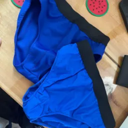 Dwie pary niebieskiej bielizny damskiej z czarną gumą, leżące na drewnianym stole obok podkładek w kształcie arbuza i czarnego portfela.