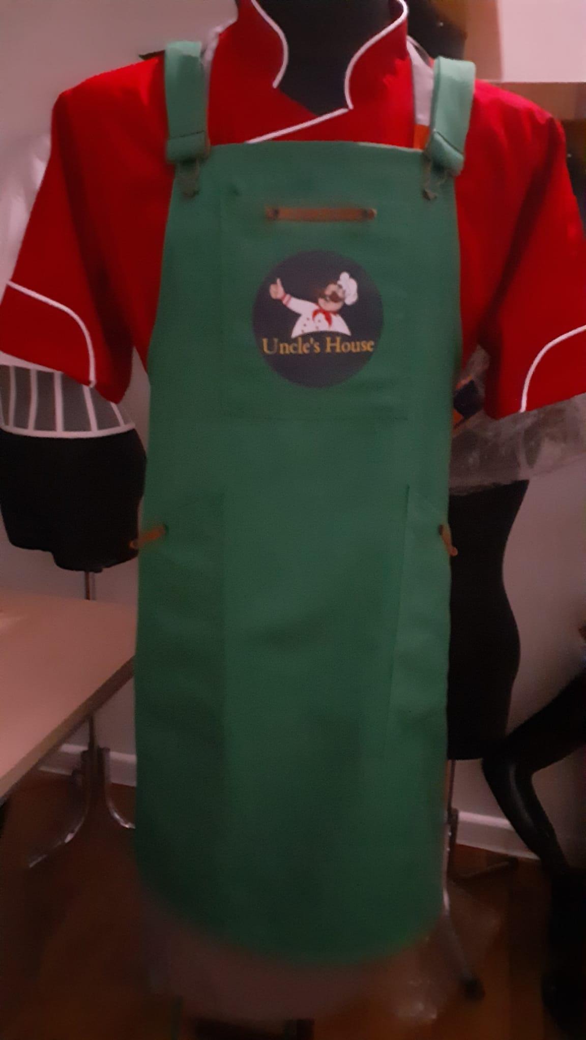 Zielony fartuch z logo 'Uncle's House' na manekinie z czerwoną kurtką kucharską, widoczne elementy stroju roboczego.