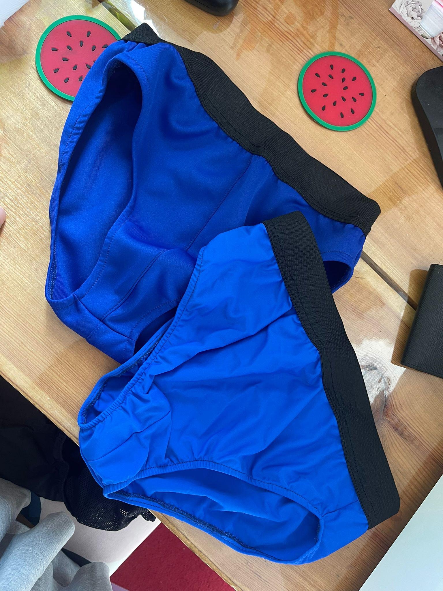 Dwie pary niebieskiej bielizny damskiej z czarną gumą, leżące na drewnianym stole obok podkładek w kształcie arbuza i czarnego portfela.