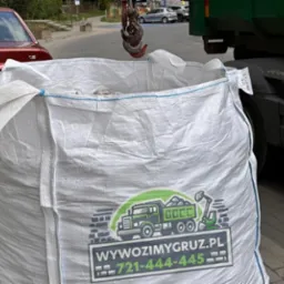 Wielki biały worek typu Big Bag z logo firmy transportowej, wypełniony gruzem, zawieszony na haku dźwigu, obok zaparkowany czerwony samochód osobowy i zielona ciężarówka.