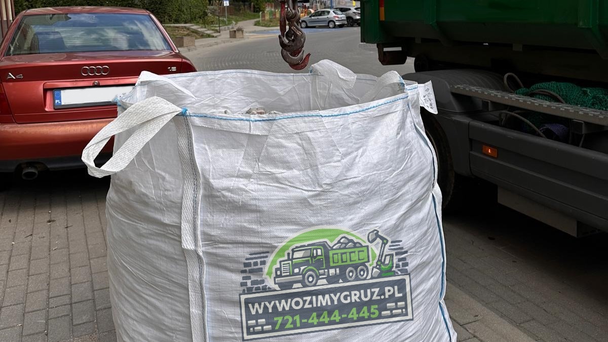 Wielki biały worek typu Big Bag z logo firmy transportowej, wypełniony gruzem, zawieszony na haku dźwigu, obok zaparkowany czerwony samochód osobowy i zielona ciężarówka.