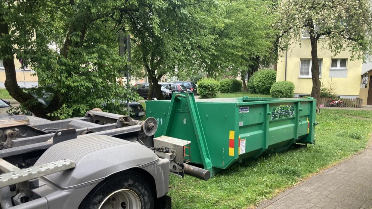 Zielony kontener na gruz umieszczony na trawie obok chodnika, podpięty do ramy ciężarówki, w tle budynek mieszkalny i drzewa.