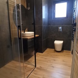 Paweł Pielak - Nowoczesna łazienka z szarą płytką ścienną, drewnopodobną podłogą, kabiną prysznicową typu walk-in z czarnymi ramami i wiszącą toaletą.