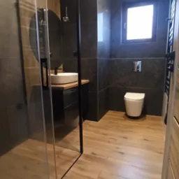 Nowoczesna łazienka z szarą płytką ścienną, drewnopodobną podłogą, kabiną prysznicową typu walk-in z czarnymi ramami i wiszącą toaletą.