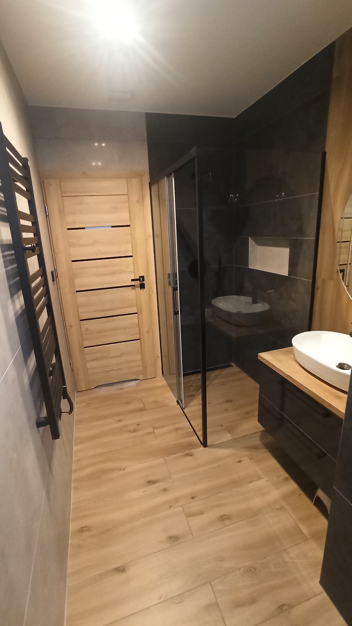 Nowoczesna łazienka z drewnianymi akcentami, kabiną prysznicową typu walk-in, grzejnikiem drabinkowym i drewnopodobną podłogą.