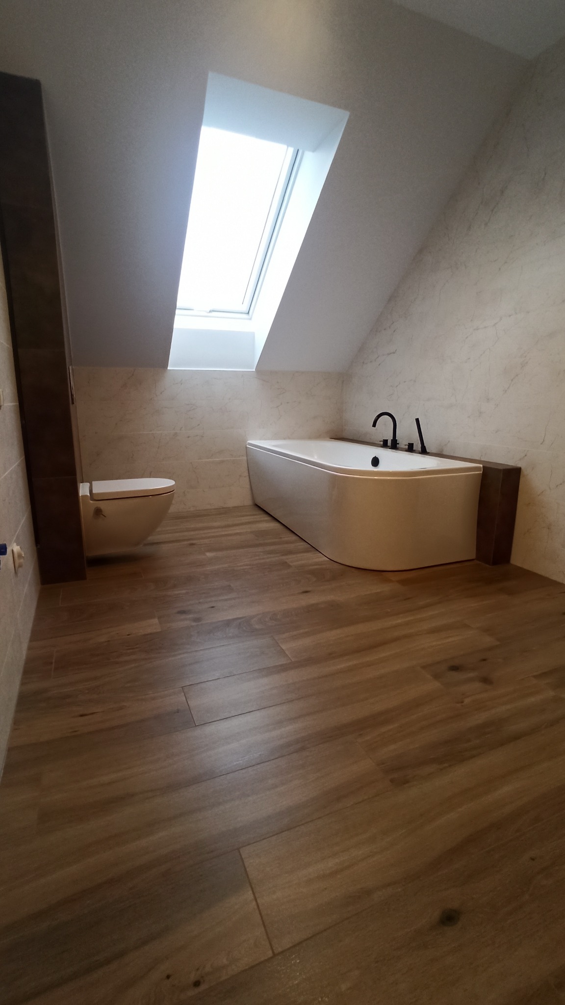 Nowoczesna łazienka na poddaszu z oknem dachowym, drewnopodobna podłoga, biała wanna z czarną armaturą i podwieszana toaleta.