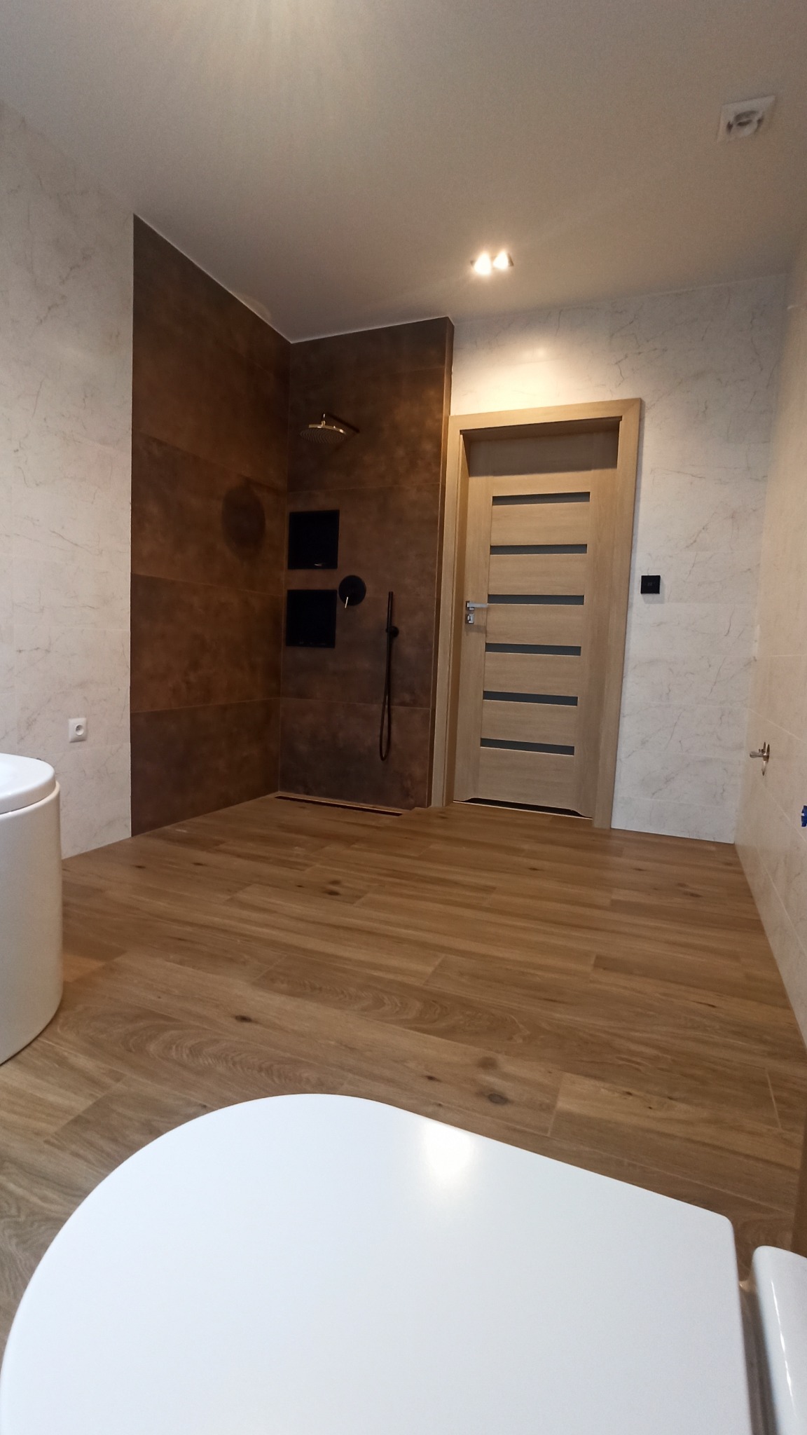 Nowoczesna łazienka z brązowymi i marmurkowymi płytkami, drewnopodobną podłogą i czarną armaturą prysznicową, częściowo widoczna toaleta z przodu.