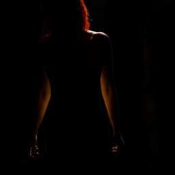 RafalSaduraFotograf.pl - Sylwetka kobiety o rudych włosach, oświetlona od tyłu, na ciemnym tle, z widocznymi ramionami i częścią pleców.
