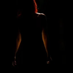 Sylwetka kobiety o rudych włosach, oświetlona od tyłu, na ciemnym tle, z widocznymi ramionami i częścią pleców.