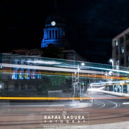 RafalSaduraFotograf.pl - Nocne ujęcie miejskiego krajobrazu z długą ekspozycją, ukazujące smugi świateł pojazdów na tle podświetlonego budynku z wieżą zegarową i nowoczesnej architektury.