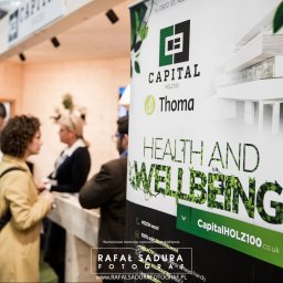 RafalSaduraFotograf.pl - Stoisko targowe firmy Capital Holz100 z grafiką promującą zdrowie i dobre samopoczucie, z widocznym logo i wizualizacją nowoczesnego budynku.