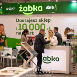 Stoisko sieci sklepów Żabka na targach, z pracownikami obsługującymi potencjalnych franczyzobiorców, na ścianie grafika z informacją o ofercie franczyzowej.